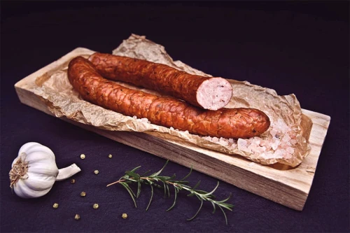 Tradycyjnie wędzona Kiełbasa Swojska od Spychalskich, prezentowana na drewnianej desce, z widocznymi ziarnami przypraw